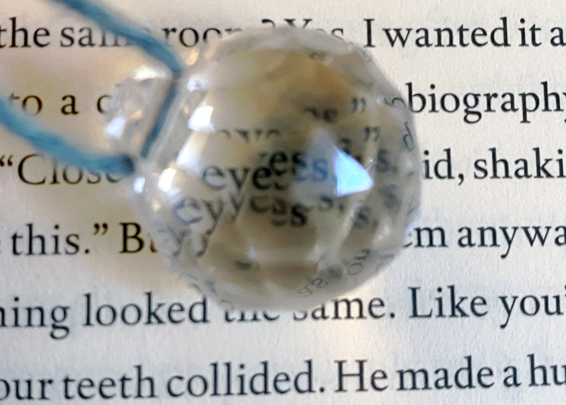 a page with printed text has a spherical crystal ontop of it. the crystal is attached to a blue piece of string also sitting ontop of the page. reflected within the crystal is the word eyes