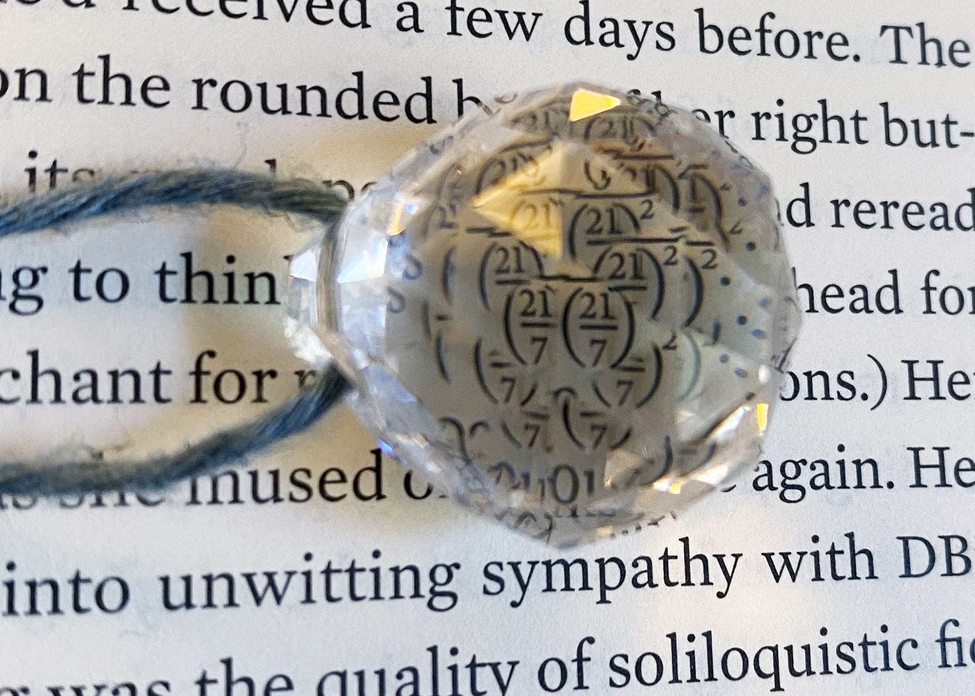 a page with printed text has a spherical crystal ontop of it. the crystal is attached to a blue piece of string also sitting ontop of the page. reflected within the crystal is the fraction (21/7)