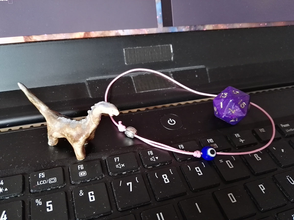 Three objects sit on the space just above a keyboard on a laptop. Arranged around the 'power' on/off button are from left to right: a small dinosaur figure, a pink string/bracelet with an evil eye on it, and a purple pearlescent 20 sided dice.
